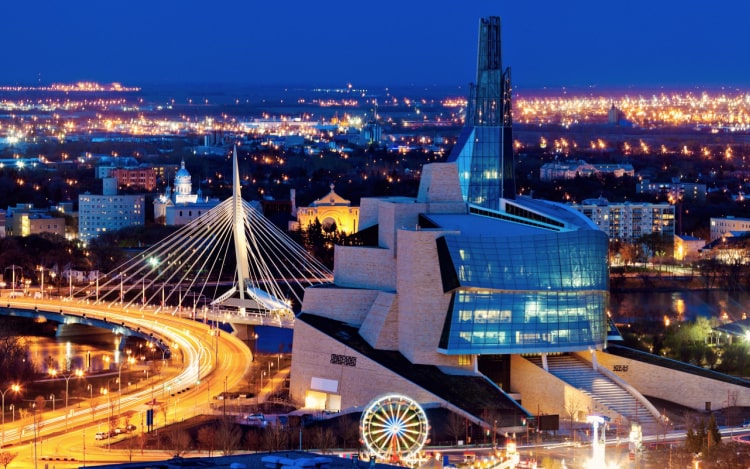 Winnipeg at night, with a large glass and grey brick building, winding road, bridge and ferris wheel all illuminated