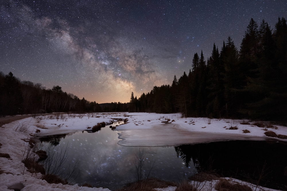 Algonquin Provincial Park at night. The Milkyway illuminates the sky above silhouettes of evergreen trees. A river winds through the foreground with snow-covered banks. The sky reflects in the water.
