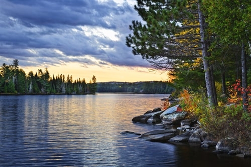 A view of Algonquin Park during sunset. The main part of the image is a view of the vast lake with forests in the distance. It is a cloudy day.