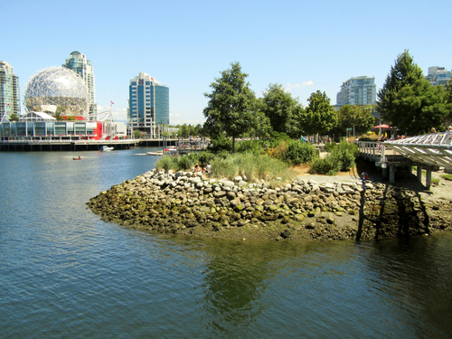 A view of a small island with a pebble beach. In the distance, you can see part of Vancouver’s city skyline.