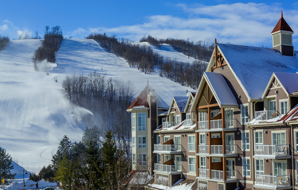 A large hotel with balconies sitting at the bottom of a ski slope. The sky is blue, and the sun is shining. Snow covers the building's roof.