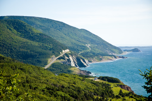 a view of the Cabot Trail along the coast of Nova Scotia. On the left, you can see forests and mountains. On the right, you can see beaches and open water.