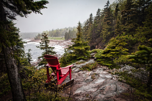 A view of the Cabot Trail from a high vantage point in the trees. There is a red deck chair in the foreground and a body of water in the background.