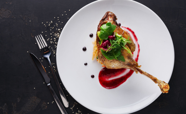 A round white plate with a cooked duck’s leg, purple-coloured sauce and green herbs on a black background, with a knife and fork resting next to the plate on the left-hand side