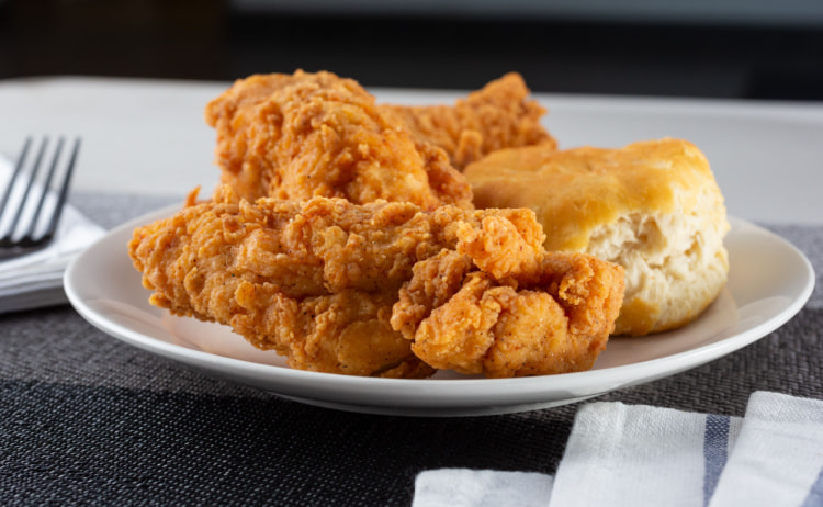 A white plate with pieces of crispy, golden-brown fried chicken and a savoury American scone sat on it. The plate is on a grey fabric placemat with a silver fork in the background.