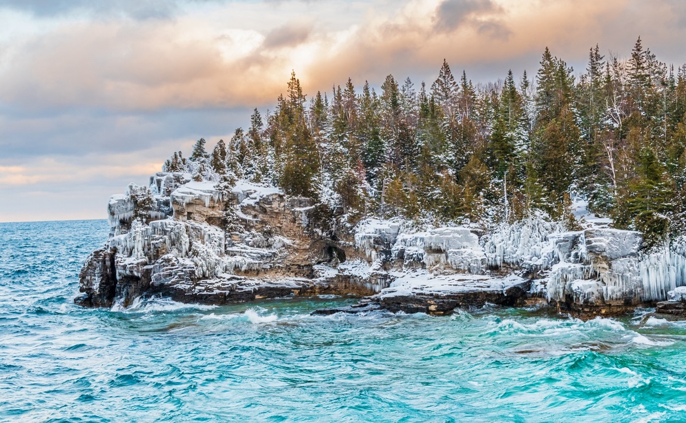 A rocky headland in Georgian Bay with snow-covered fir trees and swirling turquoise water in the lake below. The sky has peach clouds but the sun is breaking through.