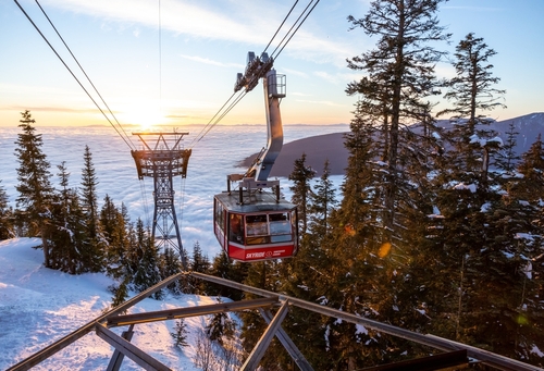 A high view of the cable cars on Grouse Mountain. You can see a red cable car approaching the peak. The sun is setting on the snowy mountain.