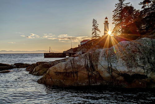 A sunset on a coastline in Vancouver. You can see a lighthouse and dock in the far distance, with trees off to the right side and the sea to the left.
