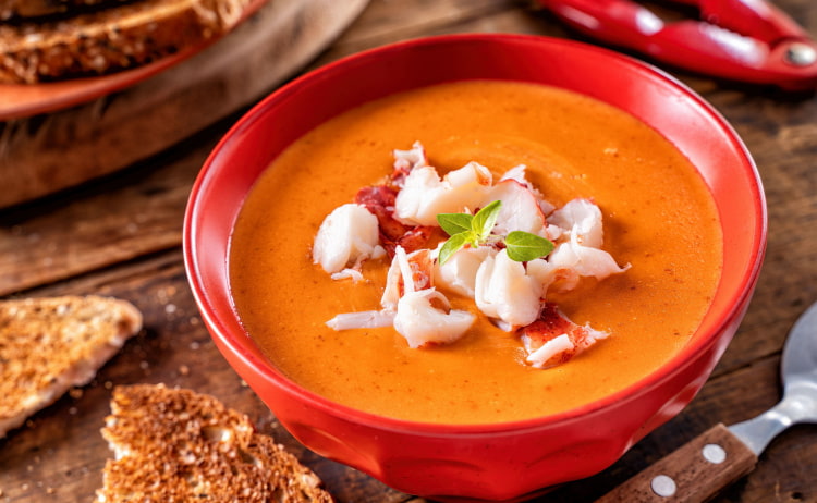 A red bowl filled with orange-coloured soup and chopped lobster, with a spoon and chunks of brown bread surrounding it