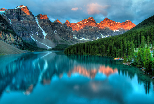 A view of Moraine Lake in Banff. The foreground shows a perfect blue lake, with a vast forest creeping in on the right side. The background is decorated with mountains that catch the sunlight.