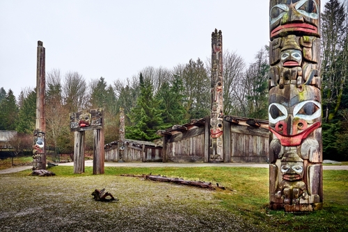 A view of an outdoor attraction at the Museum of Anthropology. The attraction features authentic wooden huts and totem poles with carvings and paintings throughout.