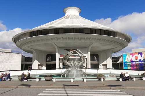 A view from the outside of the Museum of Vancouver. It is a modern-looking, white building with a water fountain and sculpture in front of it. People can be seen sitting on benches nearby.