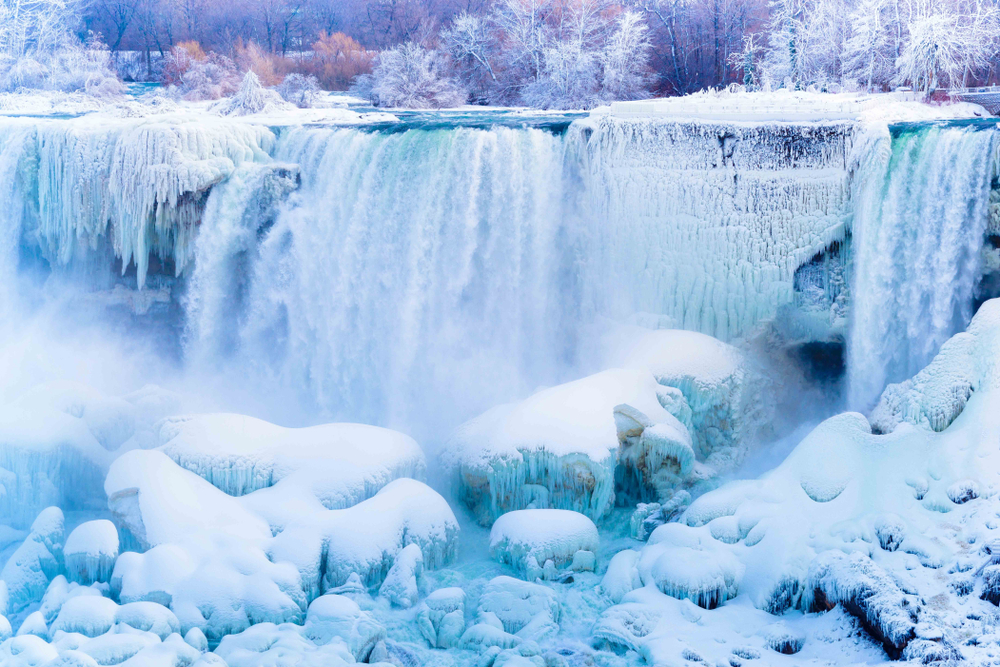 Niagara Falls frozen during the winter. The waterfall has transformed into a wall of ice, and the rocks at the bottom are covered in thick snow. Icy trees sit in the background.