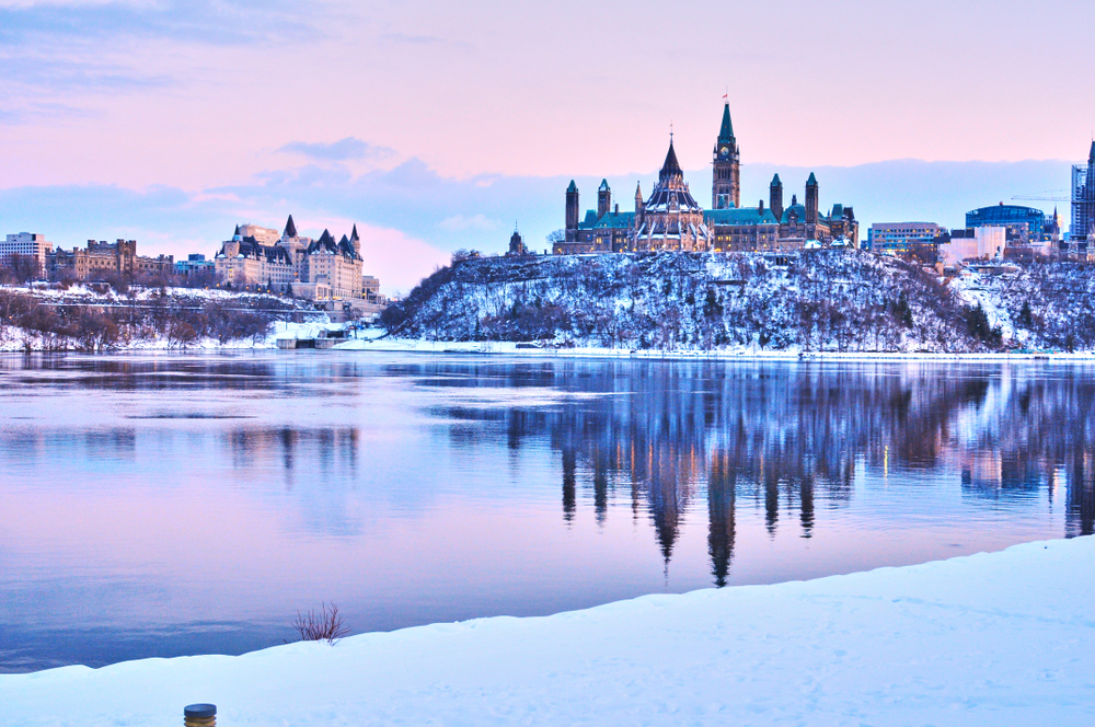 Ottawa in the winter with the city's buildings behind a semi-frozen lake and snow-covered hills. The sky is pastel pink and blue, the skyline is reflected in the still water.