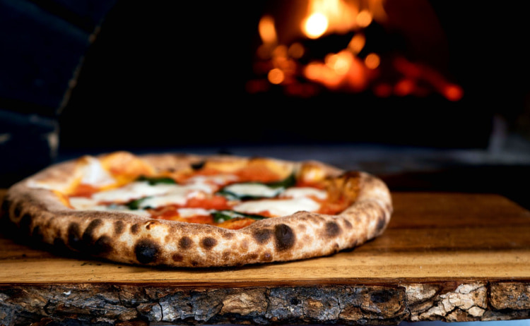 A wood-fired pizza with tomato, mozzarella and basil on top sat on a rustic wooden board with a fire just out of focus in the background.