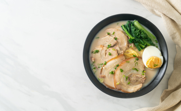 A cream-coloured broth with pieces of white meat, yellow noodles, half a boiled egg and green vegetables in it, all in a black bowl on top of a white surface.