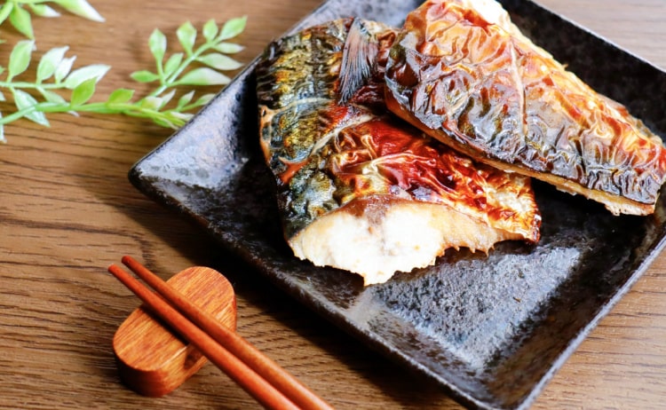 Cooked mackerel fillets with crispy skin on a square black plate, with brown chopsticks resting on the wooden surface next to the plate