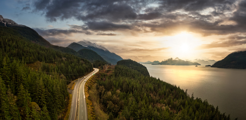 The sun setting over the Sea to Sky Highway in British columbia. From a high view, you can see the road to the left and open water and the sun setting to the right.