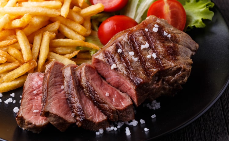 A medium-rare steak with grill lines, half-chopped into pieces and covered in rock salt sat on a black plate with french fries, chopped tomatoes and lettuce