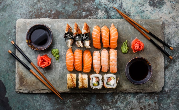 An assortment of raw fish sushi on top of a rectangular slate platter, with pieces of wasabi, ginger, and small bowls of soy sauce, as well as two pairs of brown and black chopsticks on either side of the platter