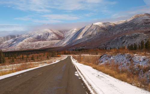 A view of a wintery road heading forward. There is autumnal, brown trees by the roadside and a grey, snowy mountain in the background.