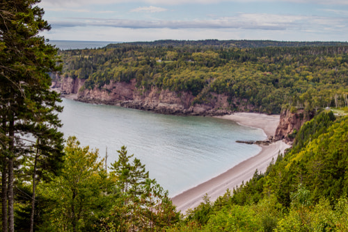 A high view of the Fundy Coastal Drive. The shot shows a bay with a small beach, some cliffs and greenery atop them.