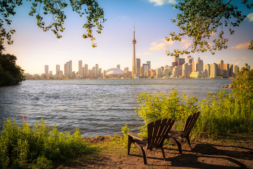 A view of Toronto from Toronto Islands. There are two deck chairs pointing towards the city from the shoreline of the island.
