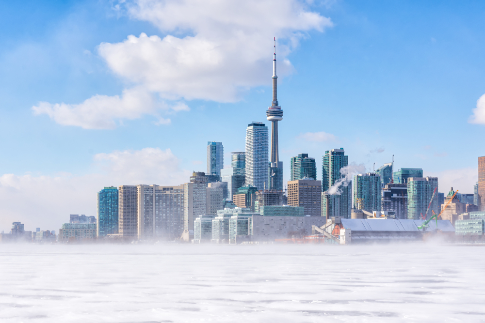 Toronto's skyline in the winter. The CN Tower and downtown buildings are sitting behind a frozen lake. The sky is blue with a few white clouds and there is a light mist on top of the ice.