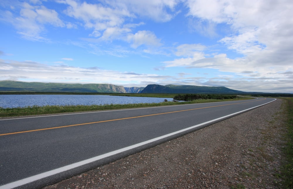 A roadside view of the Viking Trail in Newfoundland. Past the open roads, you can see a body of water. Beyond that, there are some steep mountains and cloudy skies.